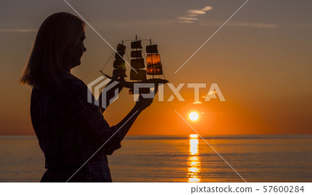 A woman stands by the sea at sunset, holds in her hands a model of a sailing ship. Hope and travel A woman stands by the sea at sunset, holds in her hands a model of a sailing ship. Hope and travel 57600284