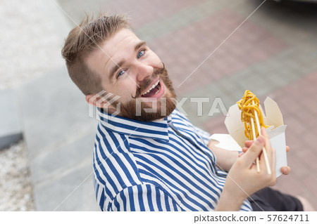 Close-up of an amusing young hipster guy eating chinese noodles with wooden chopsticks sitting in a Close-up of an amusing young hipster guy eating chinese noodles with wooden chopsticks sitting in a 57624711