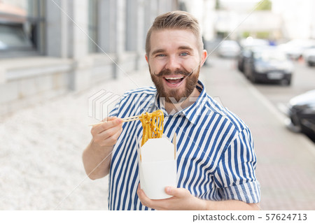 Young male hipster with a mustache and beard is eating chinese noodles with wooden chopsticks from a Young male hipster with a mustache and beard is eating chinese noodles with wooden chopsticks from a 57624713