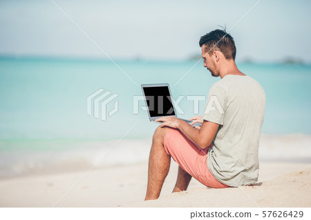 Young man sitting on sand with laptop on tropical caribbean beach. Man with computer and working on Young man sitting on sand with laptop on tropical caribbean beach. Man with computer and working on 57626429