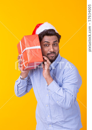 Positive young indian man Santa Claus with gifts and Christmas tree posing on a yellow background 57634005