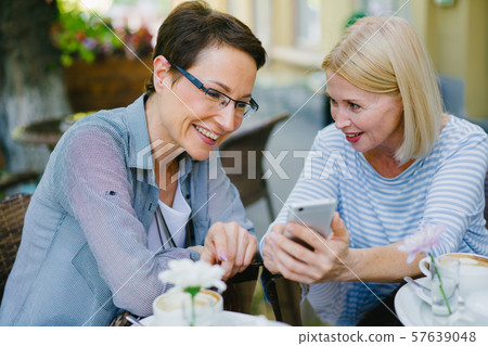 Female friends using smartphone in street cafe looking at screen having fun Female friends using smartphone in street cafe looking at screen having fun 57639048