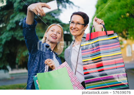 Women taking selfie with smartphone camera posing with shopping bags outdoors 57639076
