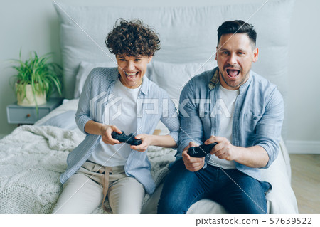 Cute couple boyfriend and girlfriend playing videogame sitting on bed in bedroom Cute couple boyfriend and girlfriend playing videogame sitting on bed in bedroom 57639522