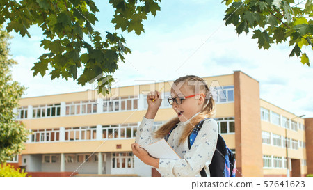 Little schoolgirl writes in a notebook against the background of her school. Little schoolgirl writes in a notebook against the background of her school. 57641623