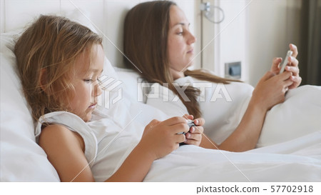 Young mother with little cute daughter are using gadgets lying on a white bed. Young mother with little cute daughter are using gadgets lying on a white bed. 57702918