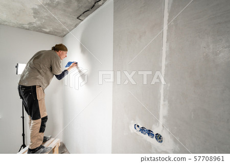 Young repairman aligns with plastic spatula. Worker glueing wallpapers on concrete wall. Repair the Young repairman aligns with plastic spatula. Worker glueing wallpapers on concrete wall. Repair the 57708961