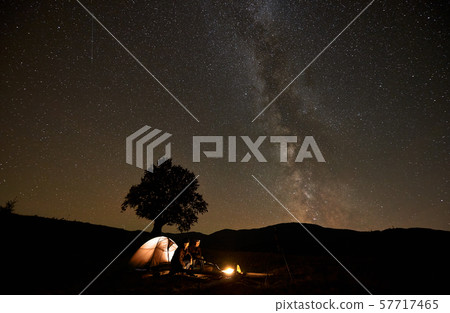 Two tourists at burning campfire in front of tent, photo camera on tripod under dark starry sky. 57717465