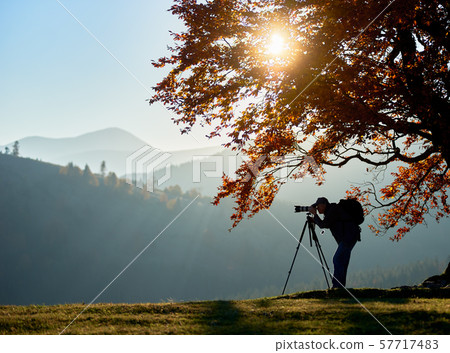 Hiker tourist man with camera on grassy valley on background of mountain landscape under big tree. 57717483