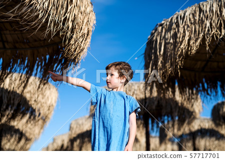 Small girl standing among straw parasols outdoors on beach. 57717701