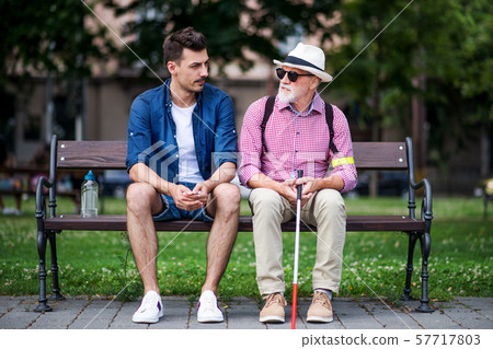 Young man and blind senior with white cane sitting on bench in park in city. Young man and blind senior with white cane sitting on bench in park in city. 57717803