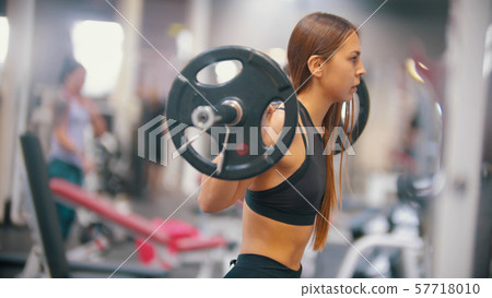 An athlete woman training in the gym - gets under the dumbbell and put it on her shoulders An athlete woman training in the gym - gets under the dumbbell and put it on her shoulders 57718010
