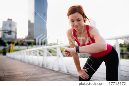 Young woman runner with earphones in city, using smartwatch. Young woman runner with earphones in city, using smartwatch. 57718572