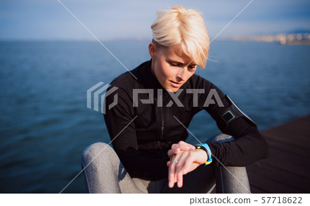 A young sportswoman sitting outdoors on beach, using smartwatch. 57718622