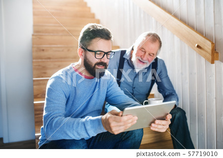 An adult son and senior father with tablet sitting on stairs indoors at home. 57719010