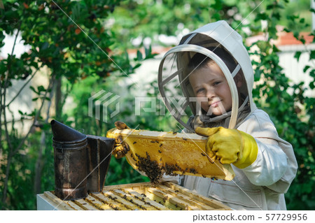 Cheerful boy beekeeper in protective suit near beehive. Honeycomb with honey. Organic food concept Cheerful boy beekeeper in protective suit near beehive. Honeycomb with honey. Organic food concept 57729956