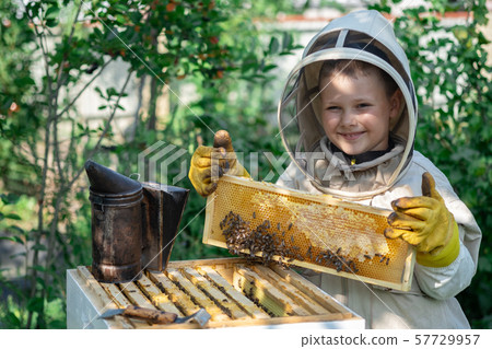 Cheerful boy beekeeper in protective suit near beehive. Honeycomb with honey. Organic food concept Cheerful boy beekeeper in protective suit near beehive. Honeycomb with honey. Organic food concept 57729957