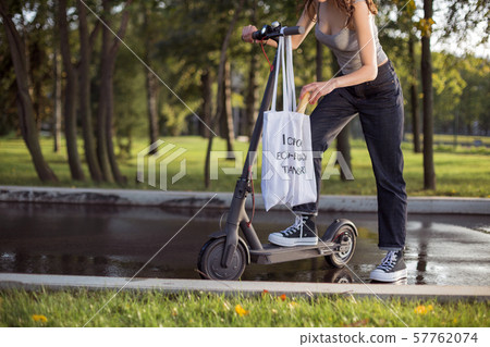 A brunette girl is standing next to electric scooter in the park and pulls out of a bag 57762074
