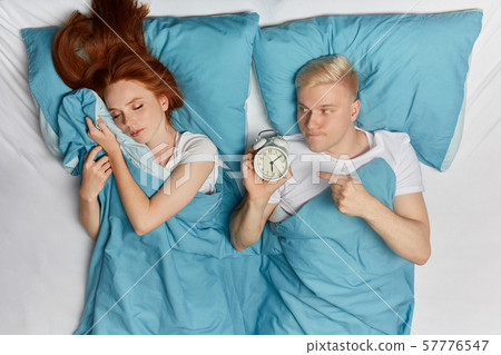 serious blonde man with alarm clock waking up young giger woman 57776547