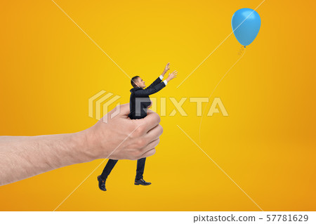 Big male hand holding tiny businessman who is reaching to blue balloon on yellow background 57781629
