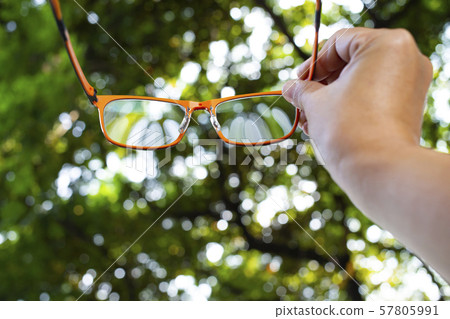 Woman's right hand holding orange eyeglasses in bokeh garden background, Close up shot, Selective focus, Image, Creative, Stationery concept Woman's right hand holding orange eyeglasses in bokeh garden background, Close up shot, Selective focus, Image, Creative, Stationery concept 57805991
