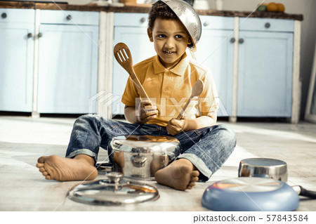 Boy sitting on the kitchen floor and playing on a saucepan while wearing a colander on the head. 57843584