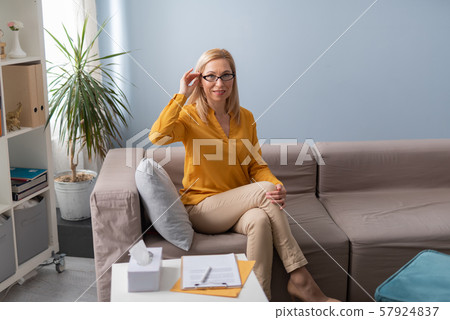 Smiling female psychologist putting on eyeglasses while sitting on sofa Smiling female psychologist putting on eyeglasses while sitting on sofa 57924837