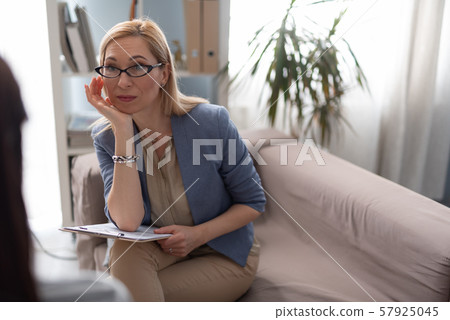 Psychologist in eyeglasses with clipboard looking attentively at patient in cabinet Psychologist in eyeglasses with clipboard looking attentively at patient in cabinet 57925045
