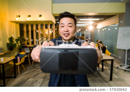 An Asian man holding virtual reality goggle on background of modern coworking studio office, his An Asian man holding virtual reality goggle on background of modern coworking studio office, his 57930042
