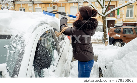 Closeup shot of preaty smiling blond lady in black coat blue jeans and hat trying to clean up snow Closeup shot of preaty smiling blond lady in black coat blue jeans and hat trying to clean up snow 57930802