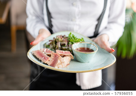 The waiter is holding a plate medium rare tuna with lettuce and hot sauce. Restaurant menu 57936055