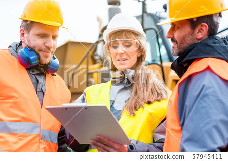 Three workers in a quarry discussing in front of heavy machinery 57945411