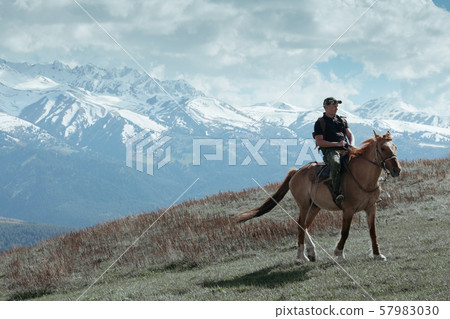 Man in cap ridding horse on field in front of mountains 57983030