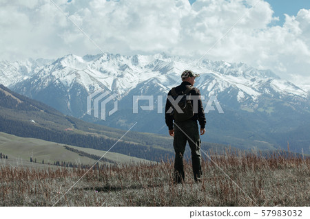Man in camouflage pants and cap standing on autumn field in front of the mountain Man in camouflage pants and cap standing on autumn field in front of the mountain 57983032