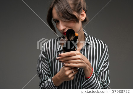 Young woman with mysterious look in striped blouse holding makeup brushes in her hands isolated 57988732