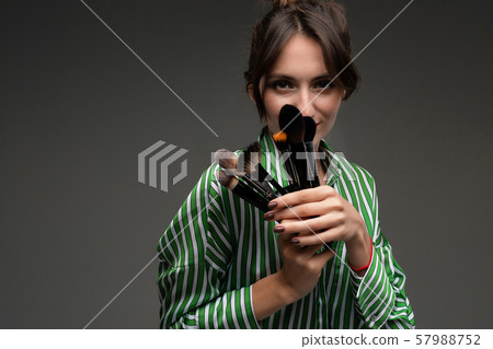 Portrait of young woman in striped blouse enjoying the softness of makeup brushes in her hands 57988752