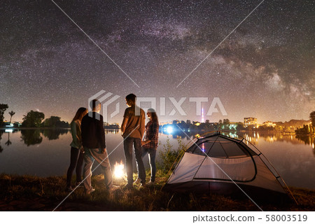 Two couple resting on lake shore near fire at night camping 58003519