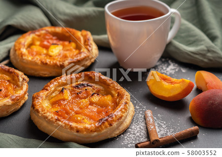 Homemade round puffs with peaches on a dark background with a linen napkin. Next to white cup with Homemade round puffs with peaches on a dark background with a linen napkin. Next to white cup with 58003552
