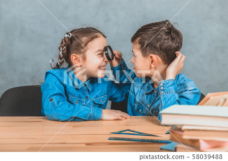 Inquisitive little schoolgirl is examining her brother with a loupe. Stack of books on the blurred Inquisitive little schoolgirl is examining her brother with a loupe. Stack of books on the blurred 58039488