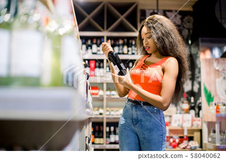 happy african american woman in pink shirt and jeans looking for wine in drink department in happy african american woman in pink shirt and jeans looking for wine in drink department in 58040062