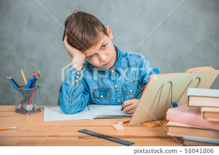 Little tired schoolboy is sitting at the desk, overwhelmed with books, leaning his head on the hand. Little tired schoolboy is sitting at the desk, overwhelmed with books, leaning his head on the hand. 58041806