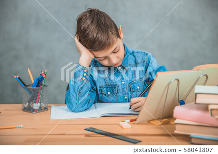 Young concentrated school kid in blue jeans t-shirt writing sitting at table full of books. Young concentrated school kid in blue jeans t-shirt writing sitting at table full of books. 58041807