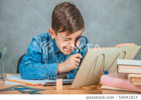 Inquisitive little boy is looking through magnifying glass at something in his book like detective. Inquisitive little boy is looking through magnifying glass at something in his book like detective. 58041813