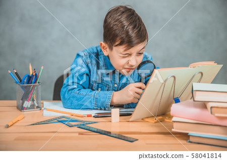 Inquisitive little boy is looking through magnifying glass at something in his book like detective. Inquisitive little boy is looking through magnifying glass at something in his book like detective. 58041814