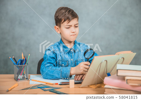 Inquisitive little boy is looking through magnifying glass at something in his book like detective. 58041815
