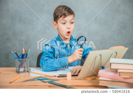 Inquisitive little boy is looking through magnifying glass at something in his book like detective. Inquisitive little boy is looking through magnifying glass at something in his book like detective. 58041816