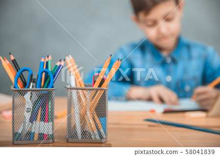 Two baskets with colored pencils, scissors and highlighters on the forefront. Schoolboy writing on Two baskets with colored pencils, scissors and highlighters on the forefront. Schoolboy writing on 58041839