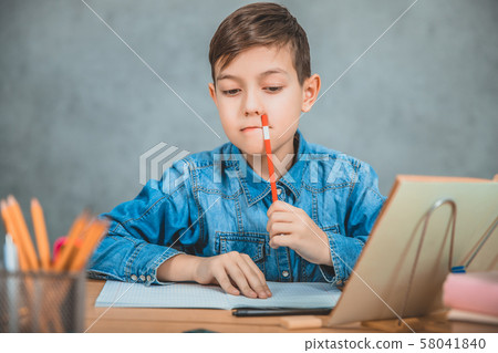 Young concentrated school kid in blue jeans t-shirt sitting at table, thinking what to write 58041840