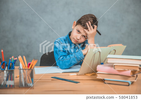 Little tired schoolboy is sitting at the desk, holding hand on the forehead, not knowing what to Little tired schoolboy is sitting at the desk, holding hand on the forehead, not knowing what to 58041853