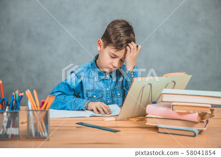 Little tired schoolboy is sitting at the desk, overwhelmed with books, holding hand on the forehead. 58041854